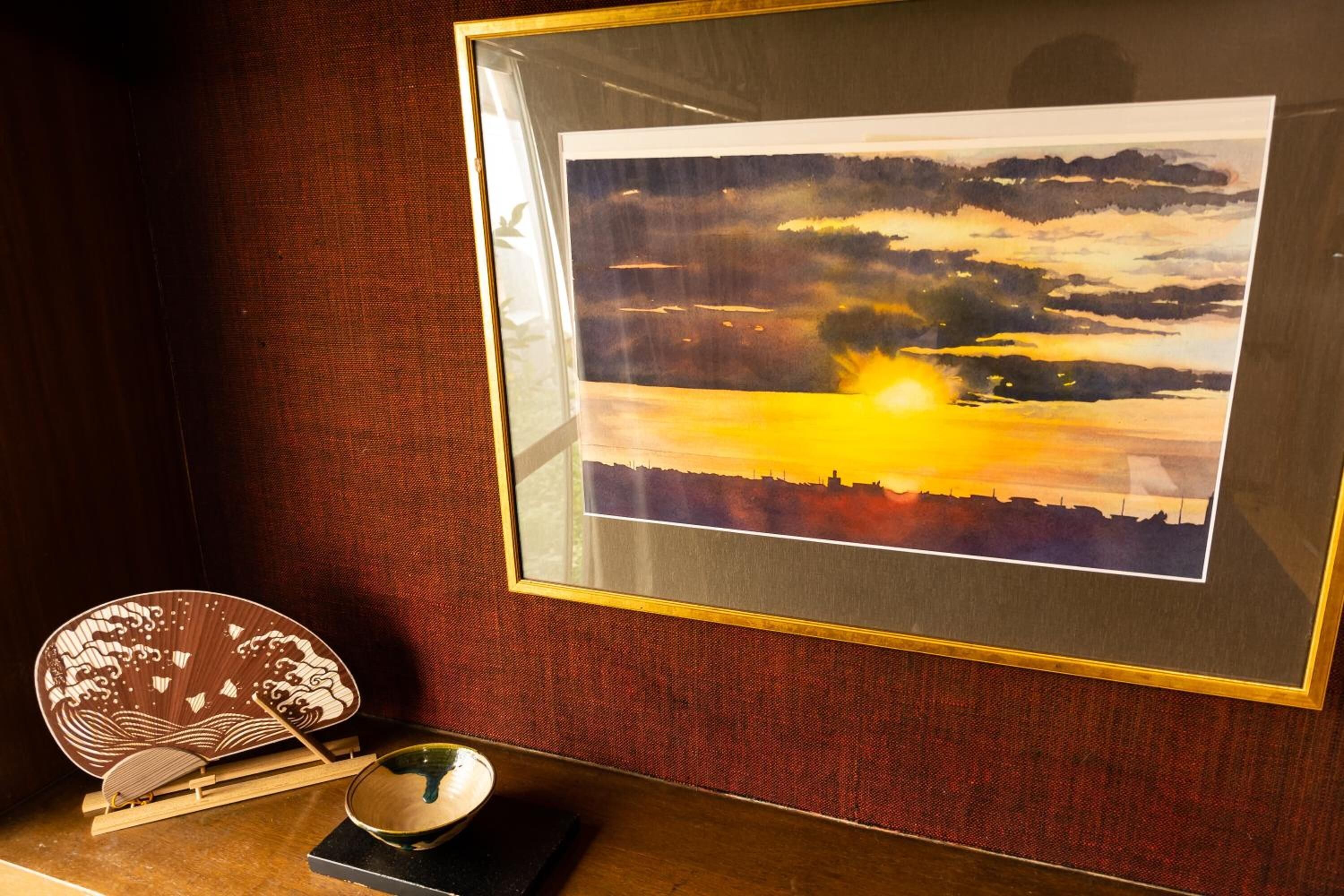 Een lijstwerk met een landschap waarop een zonsondergang afgebeeld, een traditionele Japanse waaier en een klein schaaltje op een voetstuk.