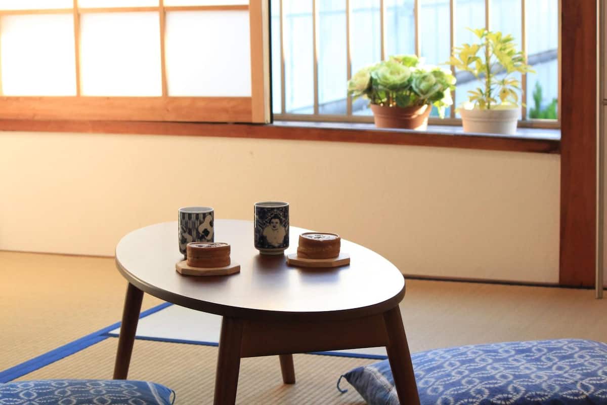 A tatami mat room with a low table, cushions, and a window with potted plants.