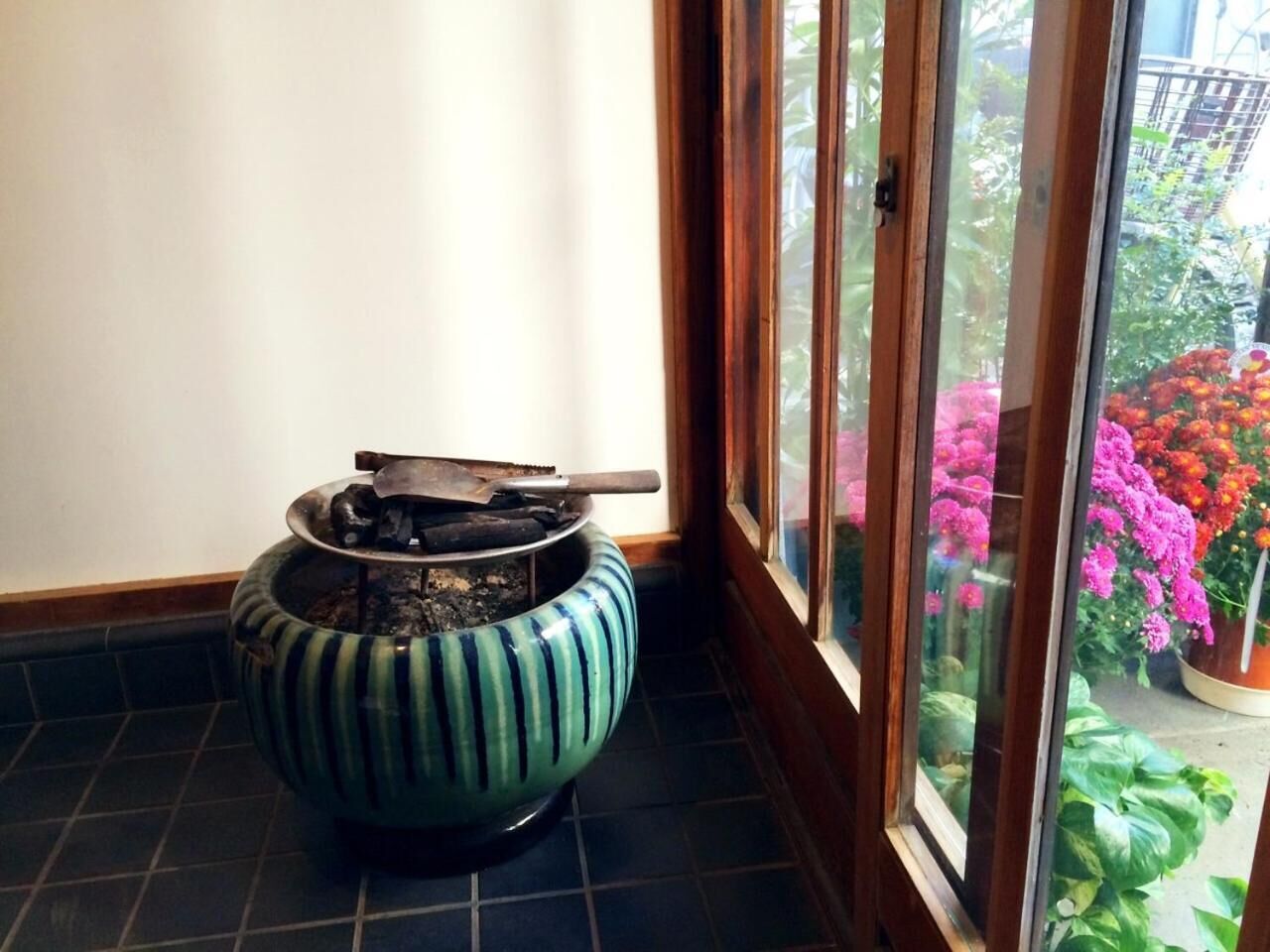 A corner with a tiled floor, a blue and green striped ceramic pot with a metal object on top, and a glass door leading to a balcony with potted plants and flowers.