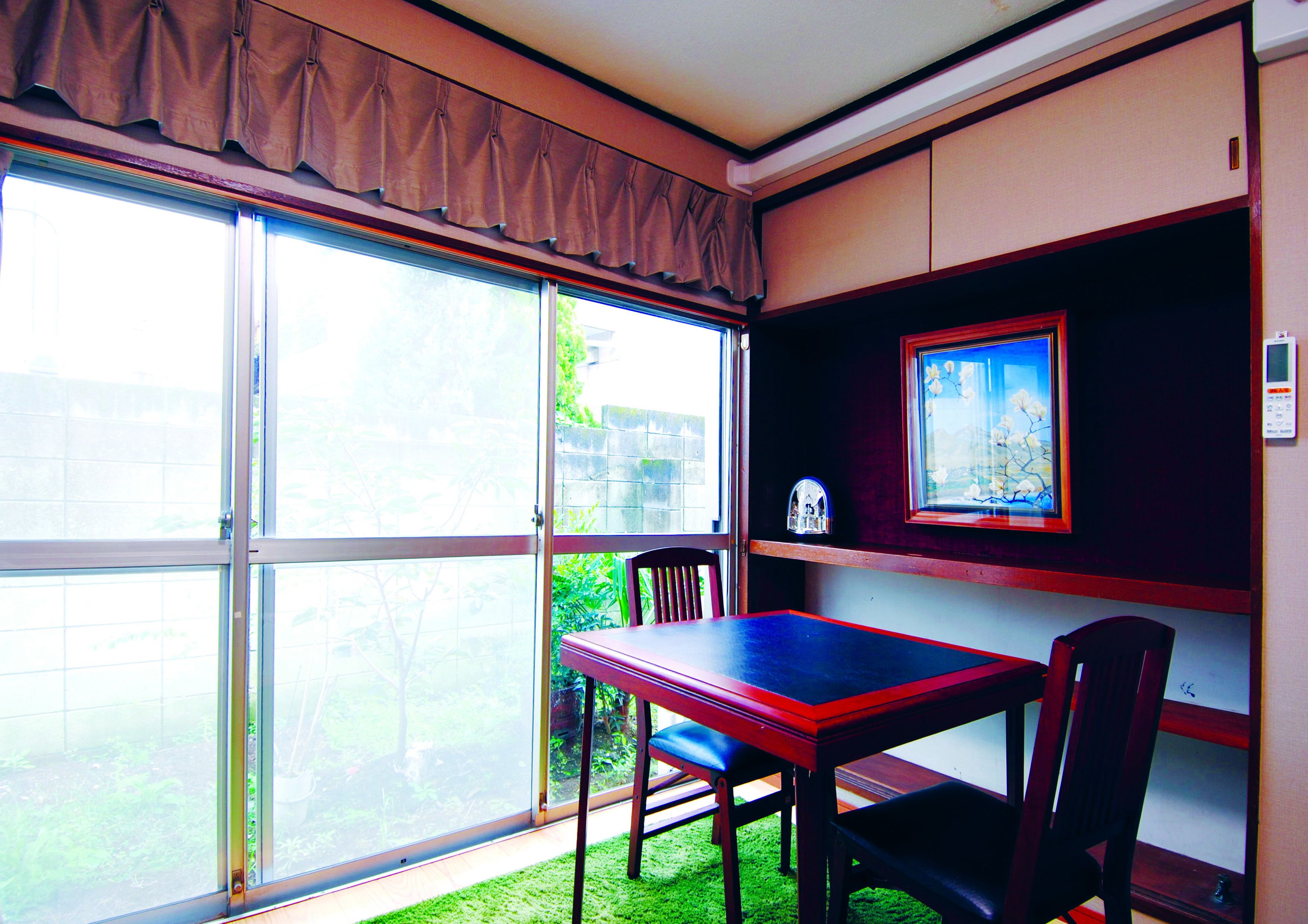 A room with a sliding glass door leading to a garden, a wooden table with chairs, a small shelf with a framed picture, and a thermostat on the wall.