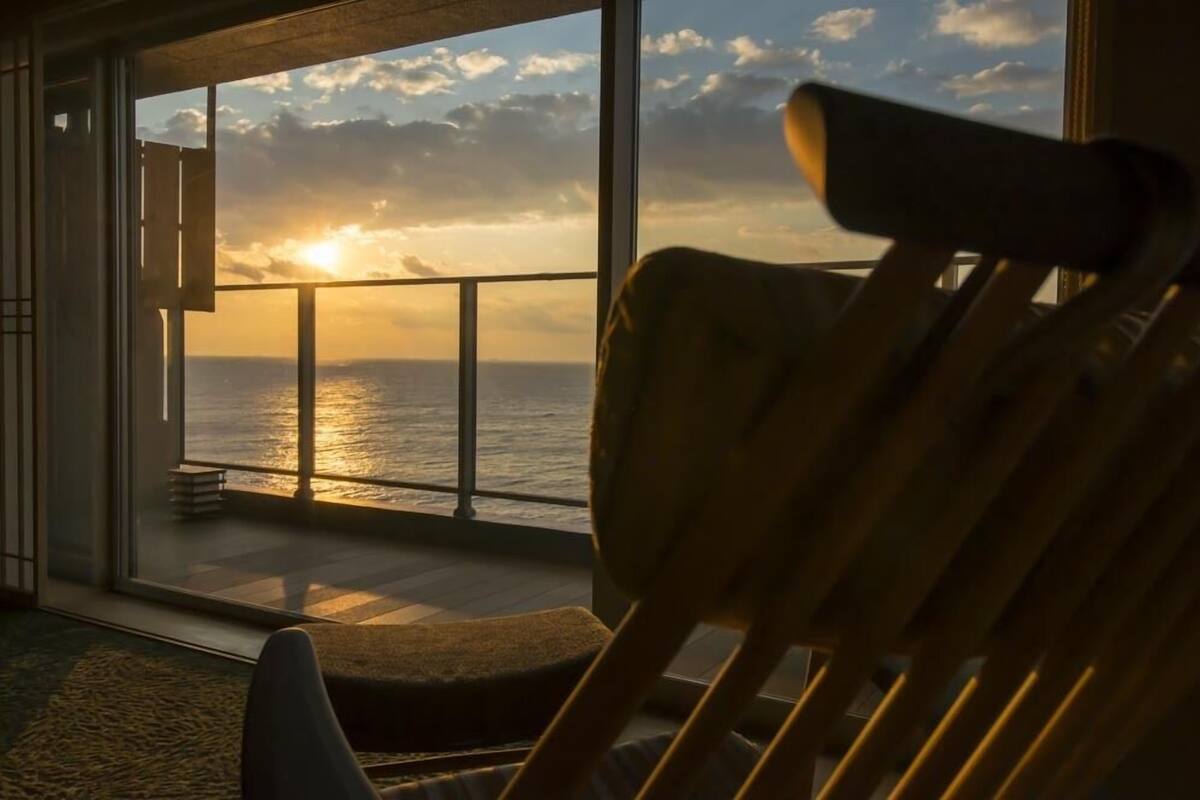 A room with a large window overlooking the ocean at sunset.
