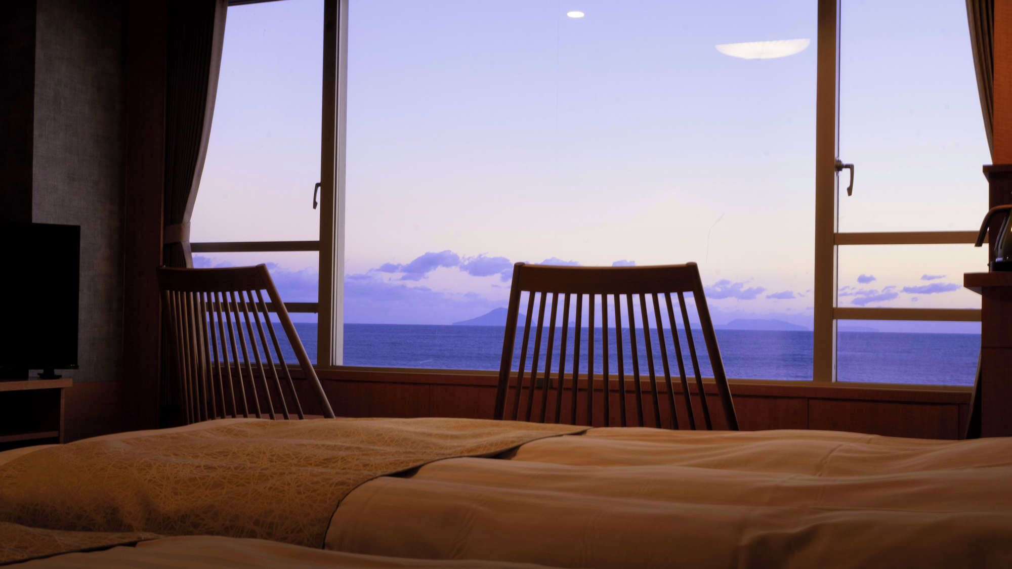 Un dormitorio con una cama, dos sillas de madera, un televisor y un ventanal con vistas al mar y a las montañas.
