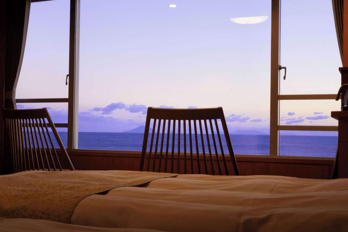 A bedroom with a bed, two wooden chairs, a television, and a large window with a view of the sea and mountains.