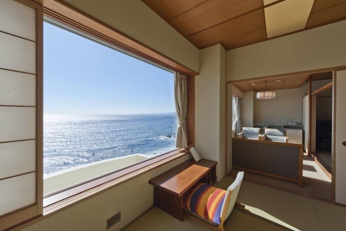 A room with a large window overlooking the ocean, a bed, a bedside table, and a small seating area with a colorful cushion.