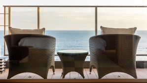 Two wicker chairs with cushions facing a glass table, overlooking a body of water through large windows. - Inatoriso (Higashiizu)