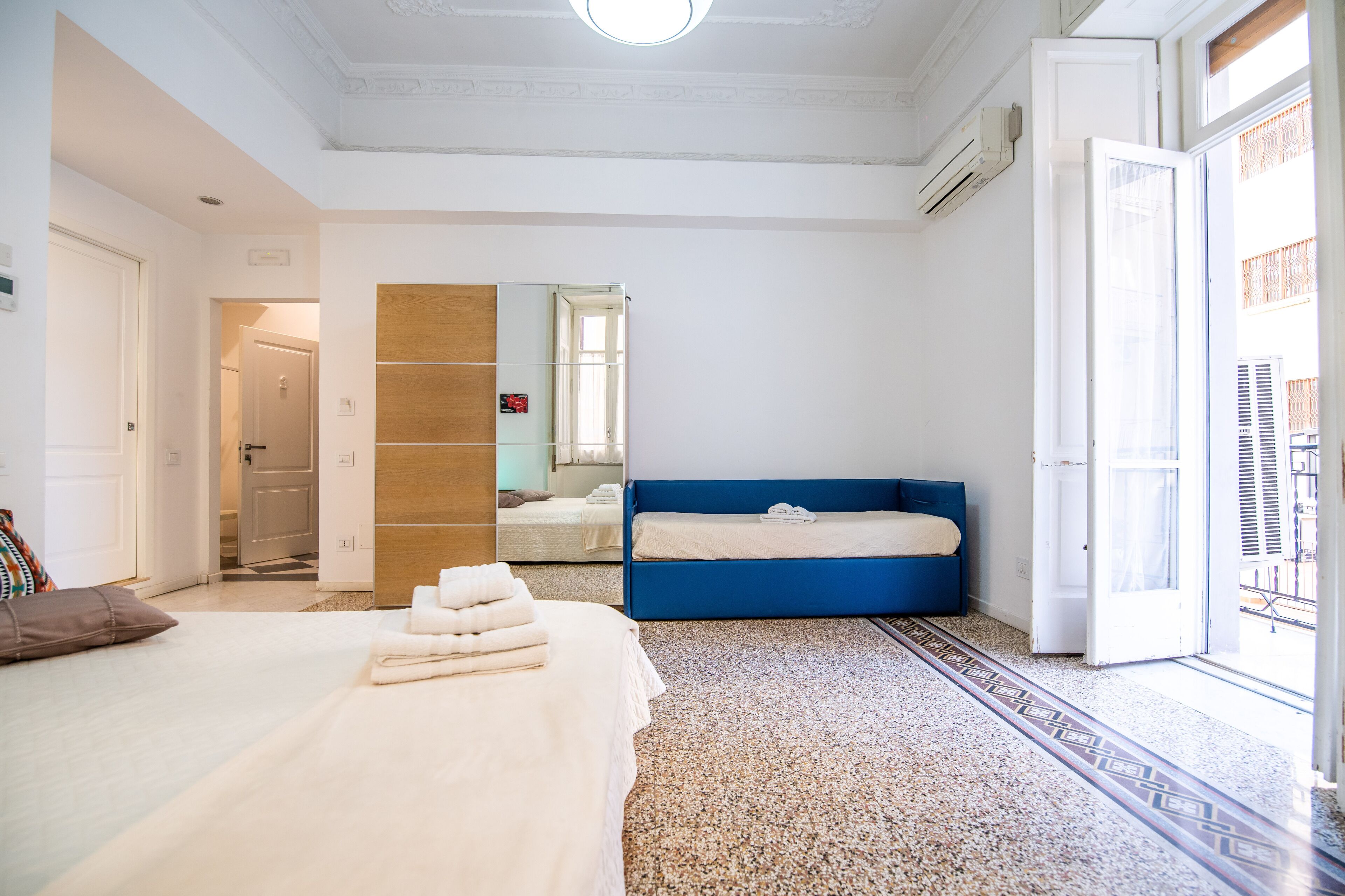 A hotel room with a bed, a blue bench, a mirror, and a door leading to a balcony.