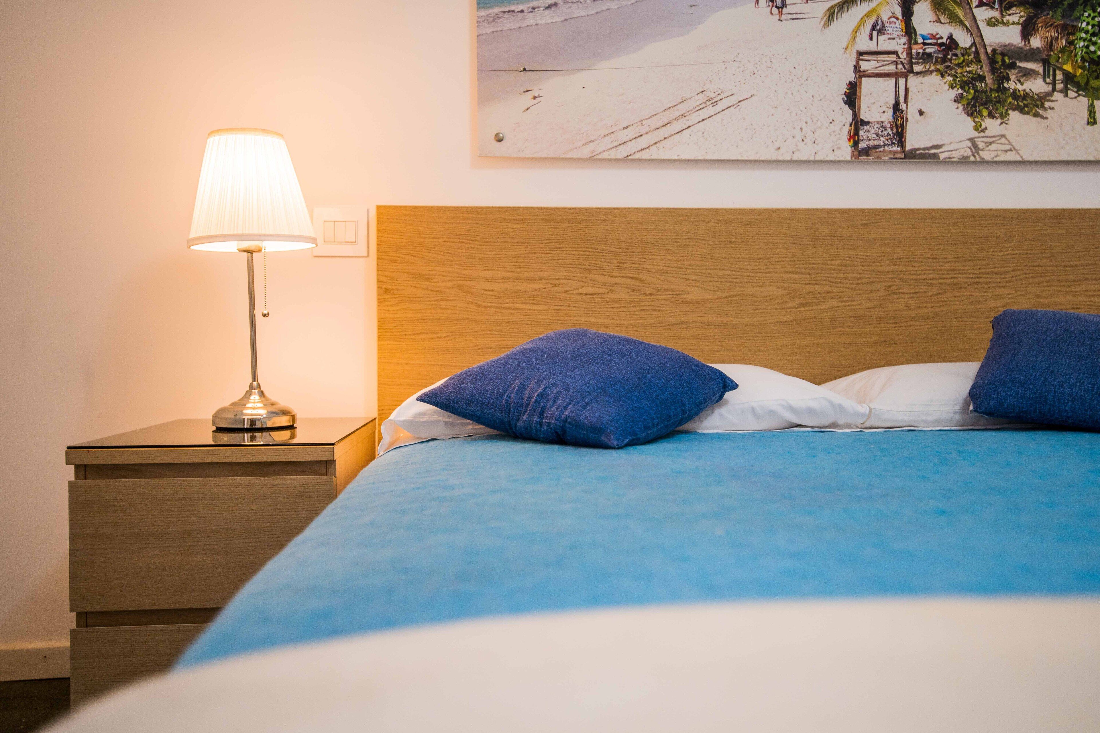 A neatly made bed with blue bedding, a wooden headboard, and a bedside lamp on a wooden nightstand.