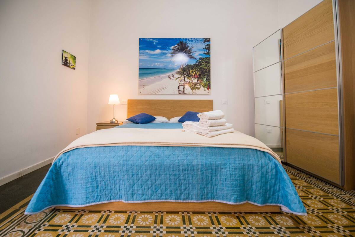 Un lit bien fait, recouvert d’une couvre-lit bleue, avec une tête de lit et un tableau représentant une scène de plage accroché au mur.