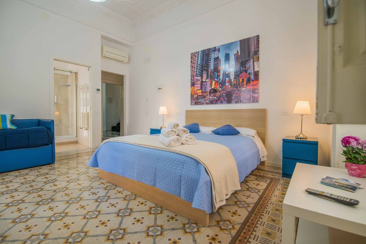 Une chambre d’hôtel avec un lit, un canapé bleu, une petite table et un tableau représentant un paysage urbain.