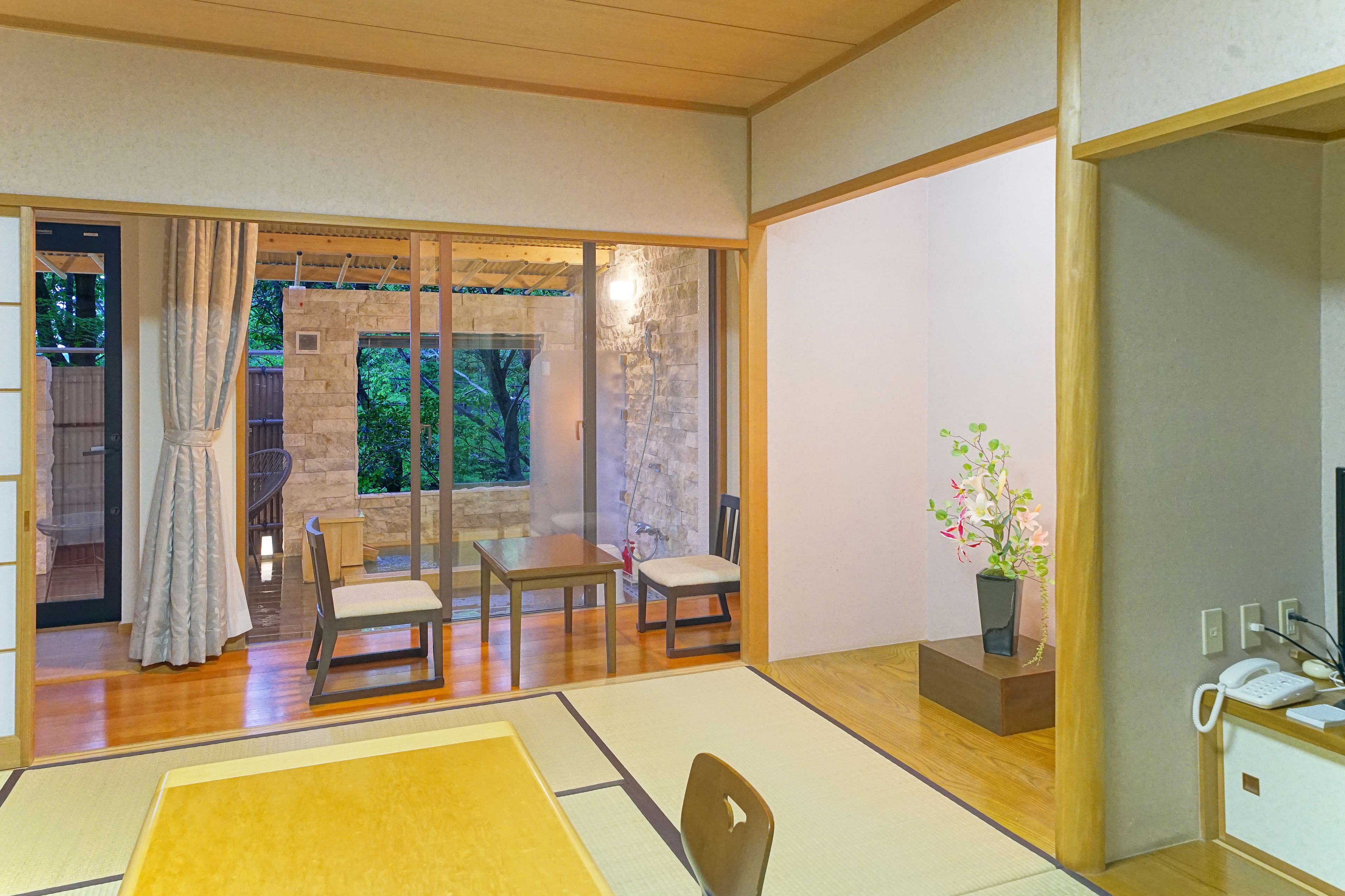 A traditional Japanese-style room with tatami flooring, a sliding glass door leading to a garden, a small table with chairs, and a potted plant.