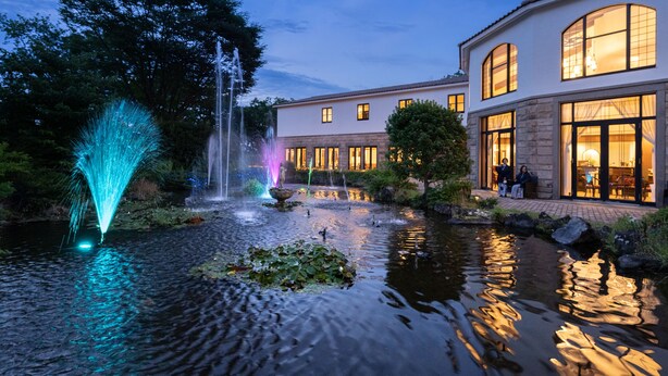 A serene pond with fountains and a well-lit building with large windows. - Mori No Izumi (Ito)