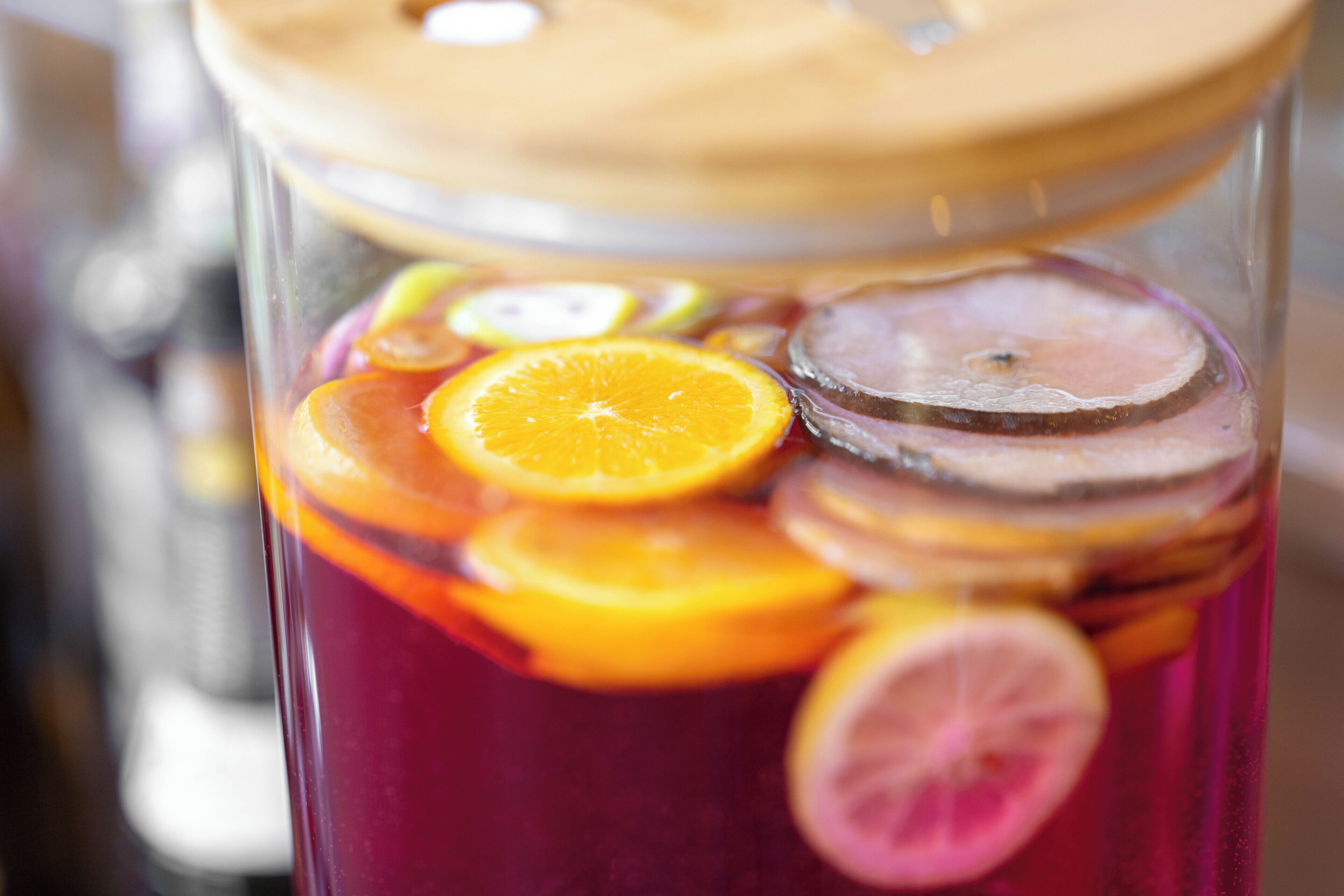 Un récipient en verre doté d’un couvercle en bois, contenant un liquide rouge et des tranches d’orange et de citron.