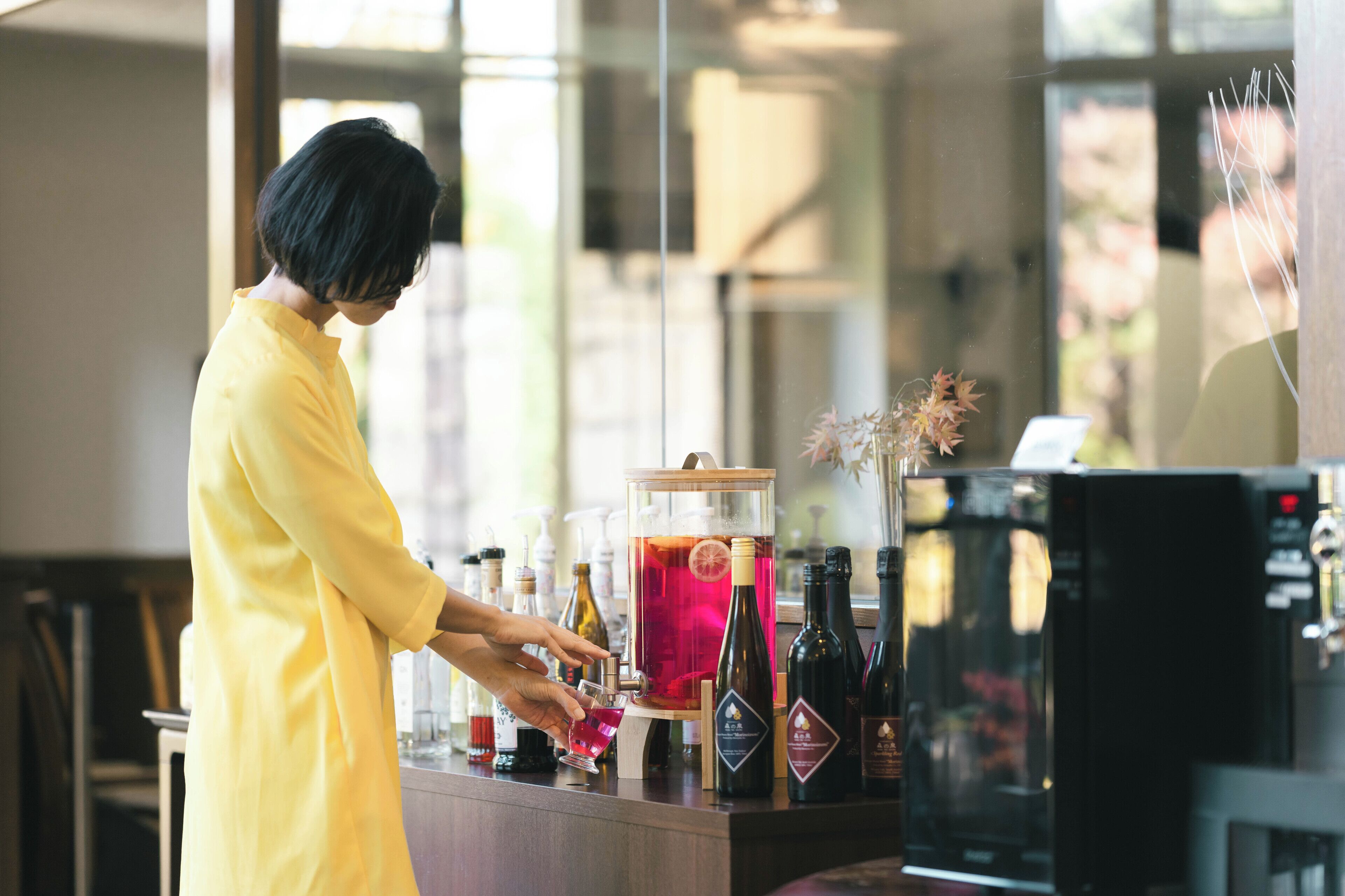 A person in a yellow dress preparing a drink at a bar with various bottles and a large glass container.