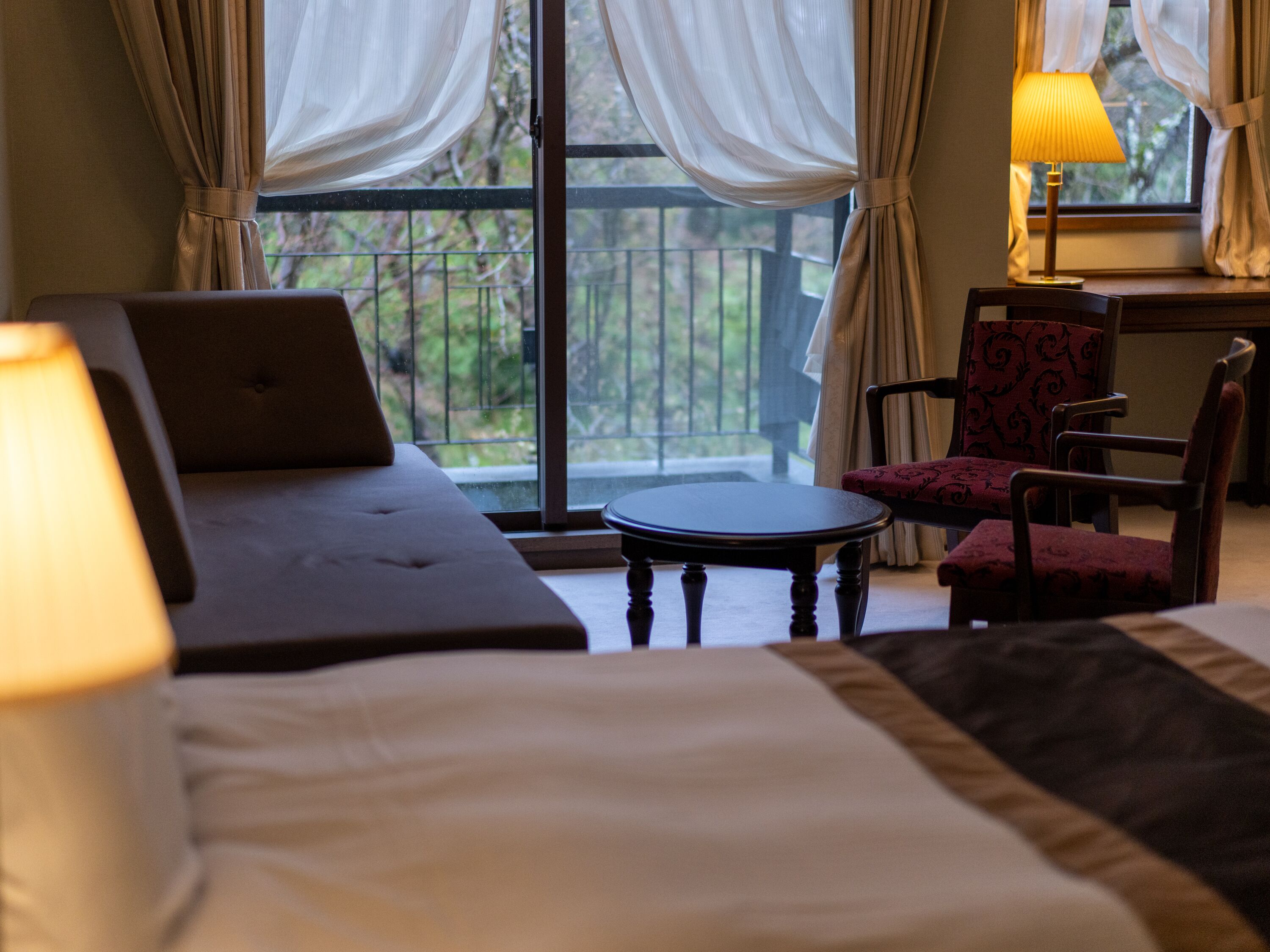 Una habitación de hotel con una cama, un ordenador portátil sobre la cama, una mesita redonda pequeña, una silla y una lámpara.