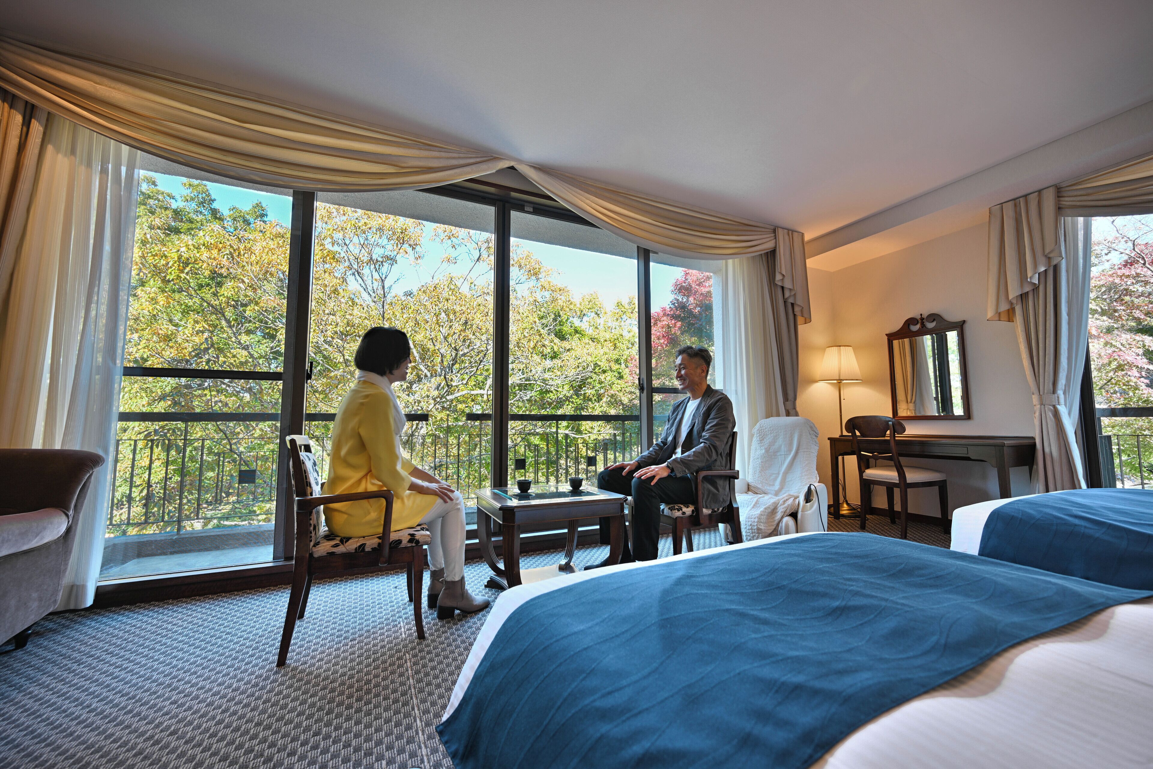 Habitación de hotel con dos camas, un escritorio y una silla. Amplia ventana con cortinas y vistas a árboles.