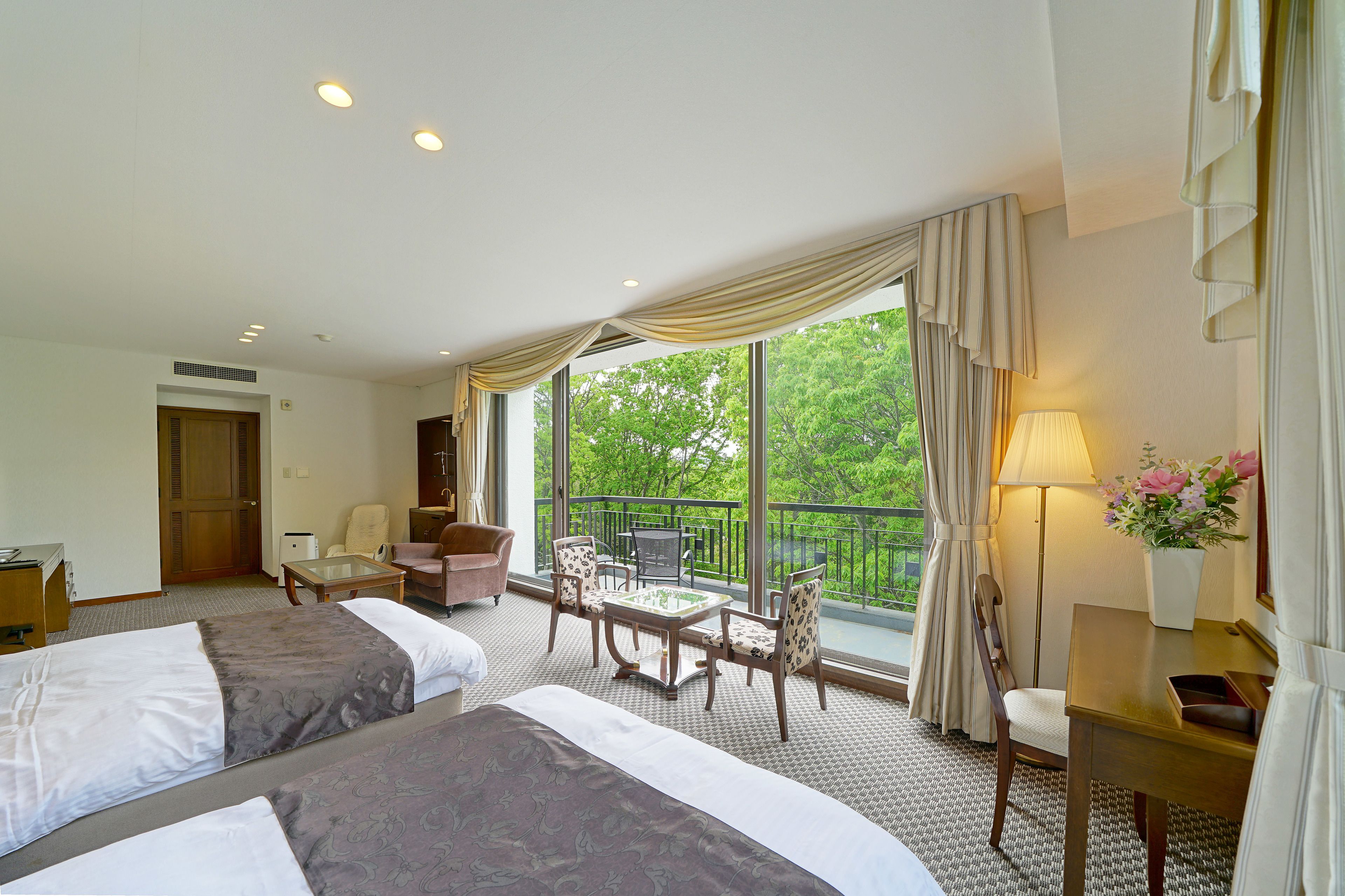 A hotel room with two beds, a balcony with a table and chairs, a desk with a lamp, and a view of greenery outside.