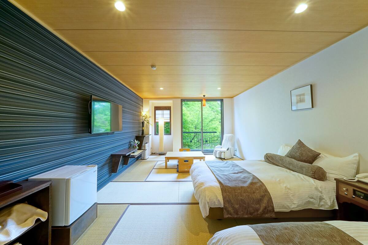 A modern hotel room with a large bed, a desk, and a view of greenery through the window.