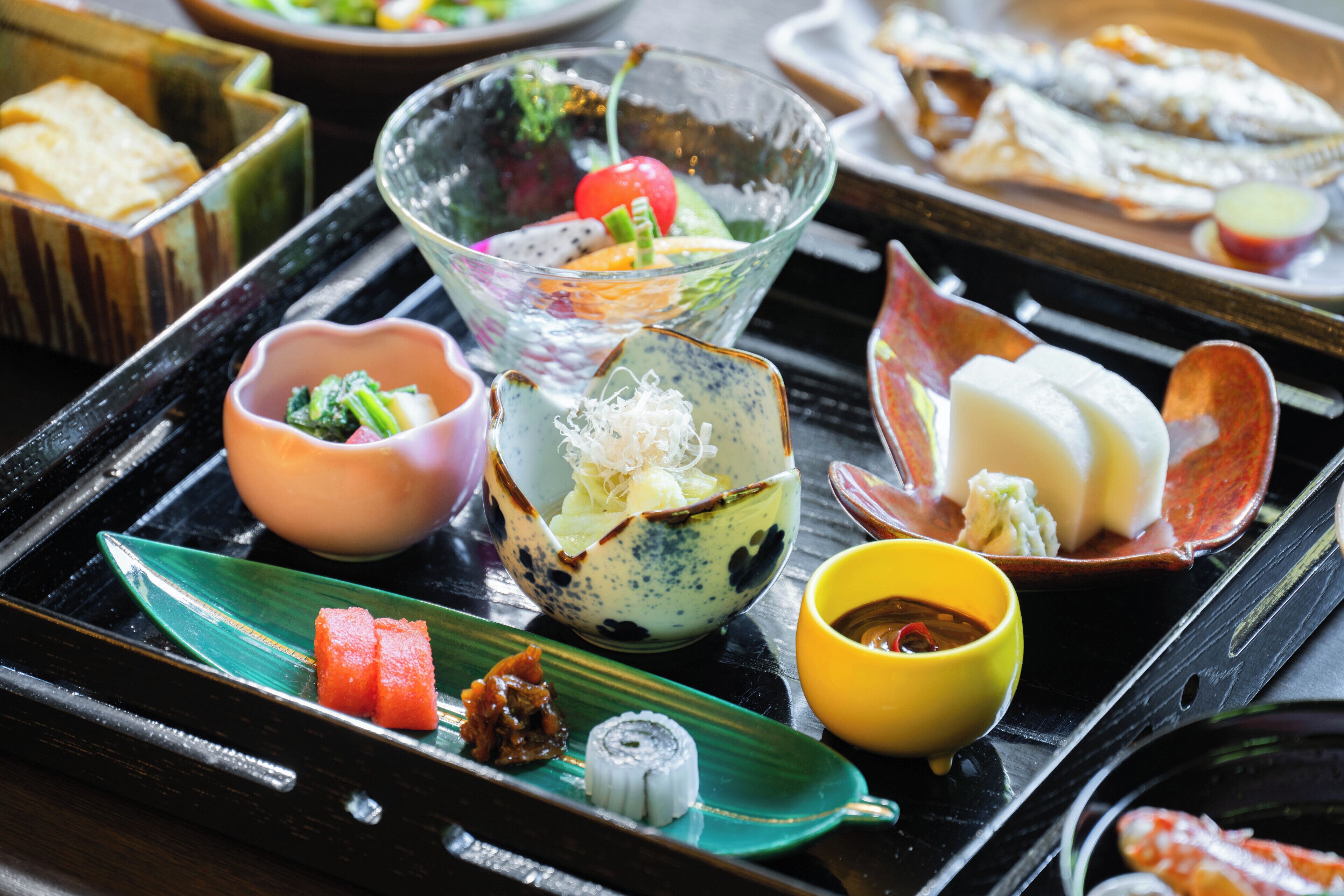 A variety of dishes on a black tray, including a bowl with a cherry tomato, a bowl with noodles, and a plate with a fish.