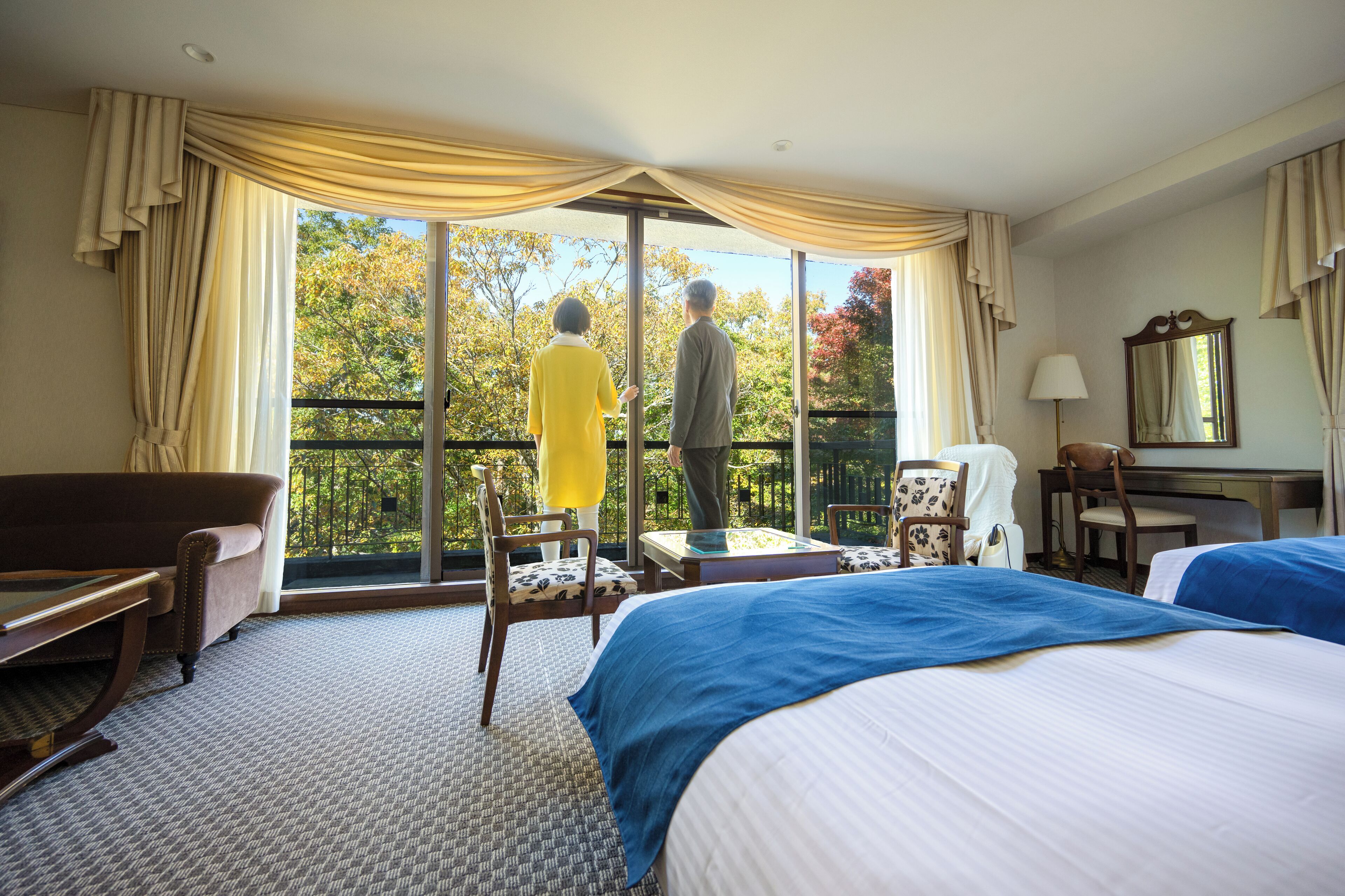 A hotel room with a large window, a sofa, two beds, a desk, and a chair.