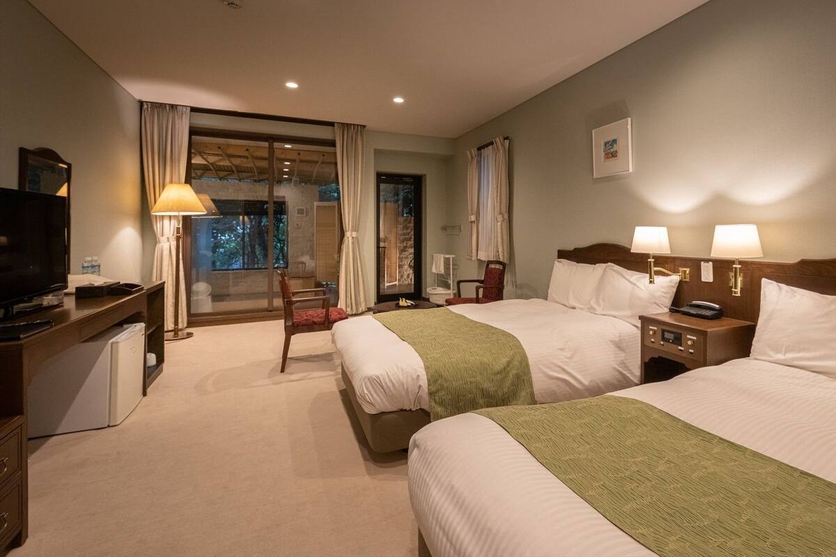 A hotel room with two beds, a desk with a TV, a chair, a nightstand with lamps, and a window with curtains.