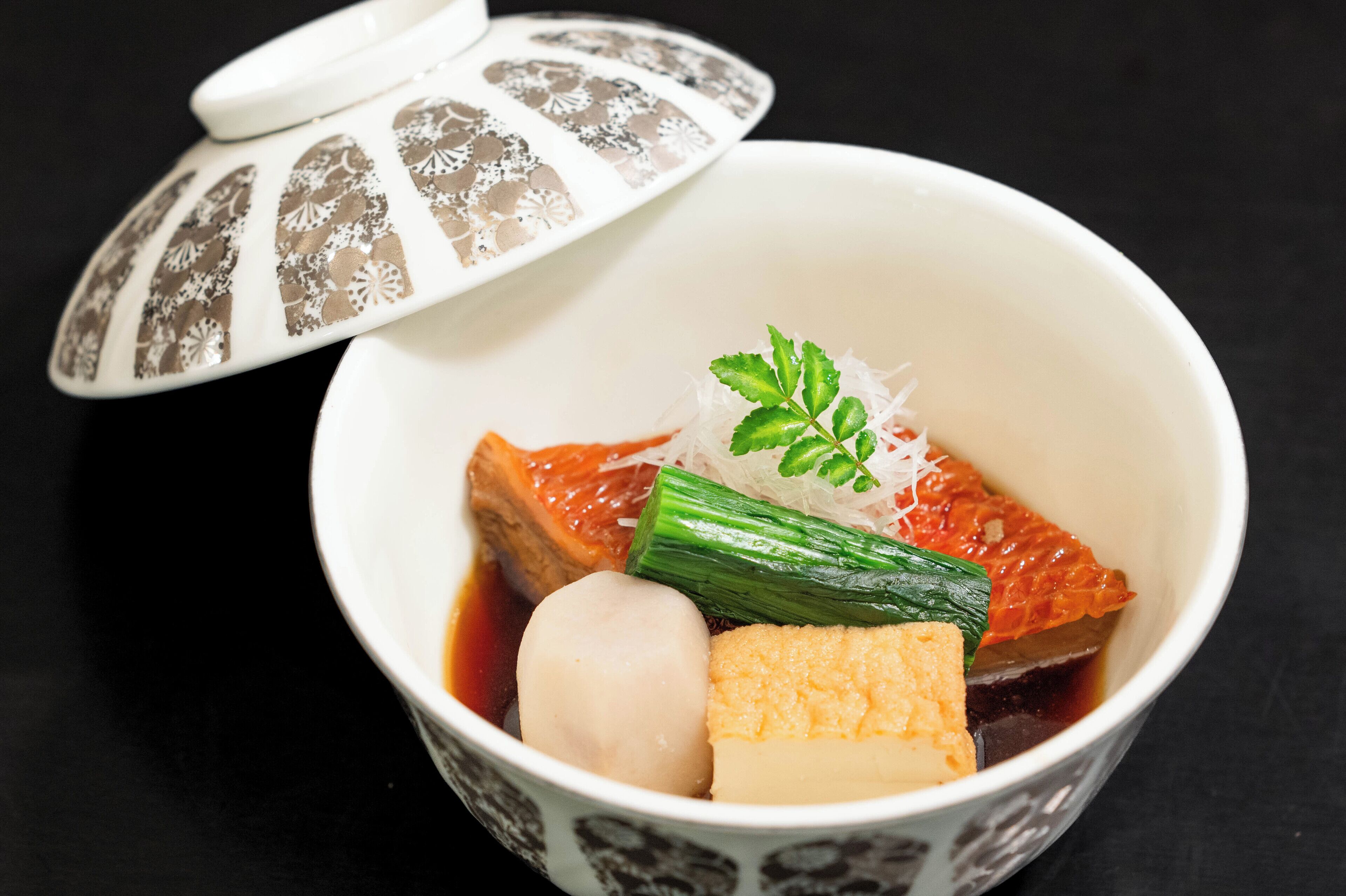 A bowl of Japanese hot pot with tofu, green onions, and a piece of fish, served with a decorative lid.