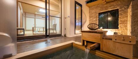 A traditional Japanese onsen with a wooden bench, a stone wall, and a window overlooking a room with tatami mats.