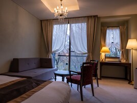 Una habitación de hotel con cama, sofá, escritorio, silla, lámpara y ventana con cortinas.