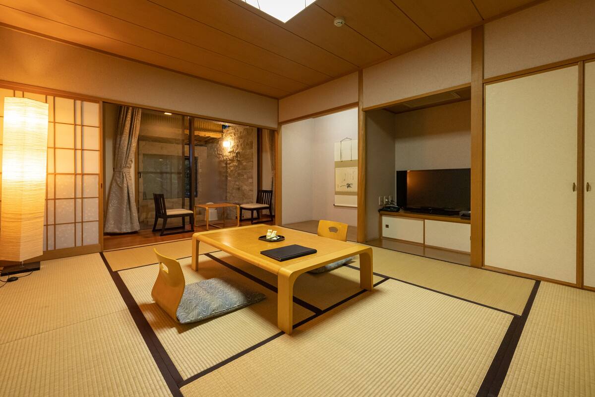 A traditional Japanese room with tatami flooring, a low table with cushions, sliding doors, and a television.