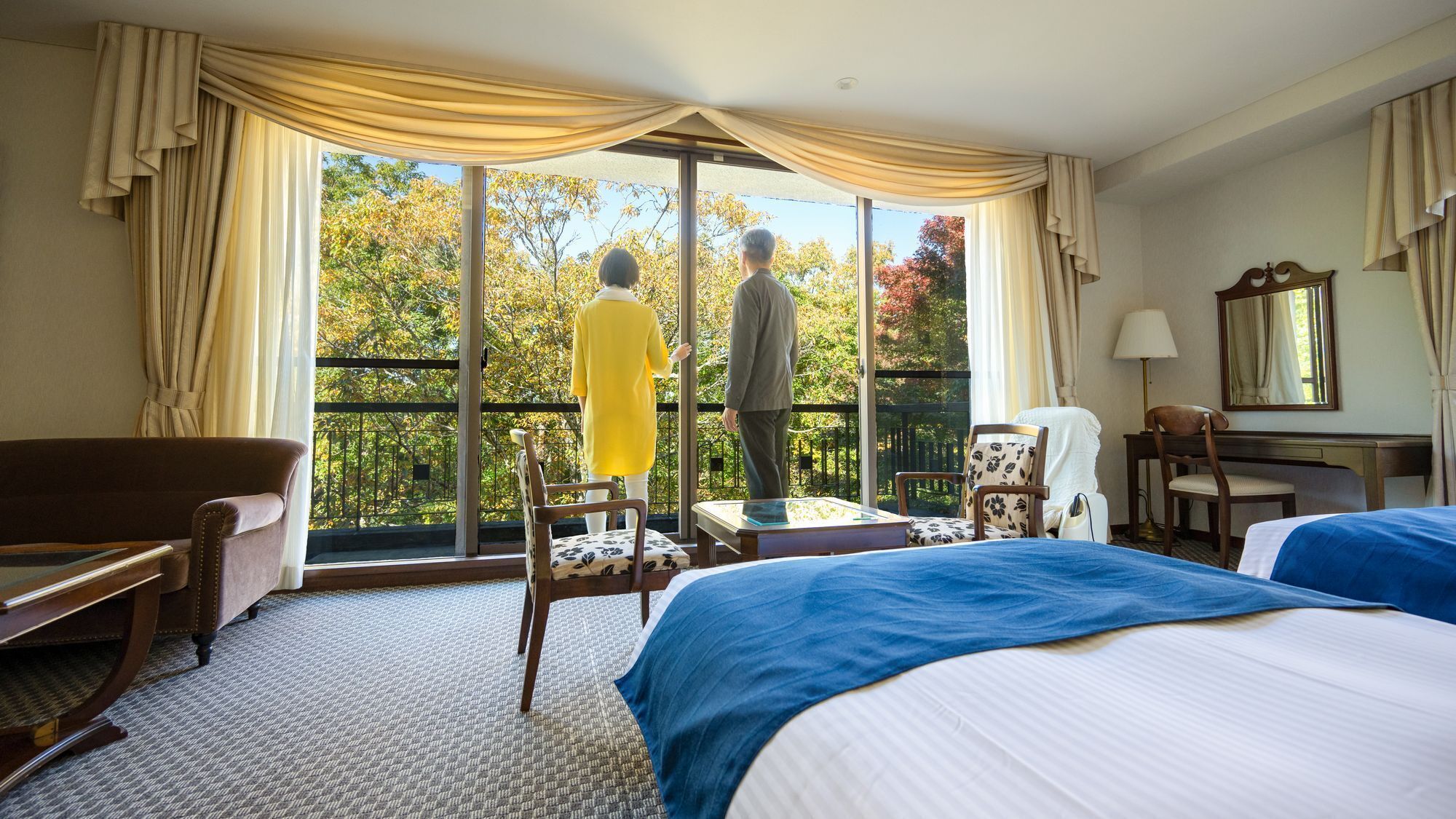 A hotel room with a large window, a bed, a desk, a chair, and a sofa.