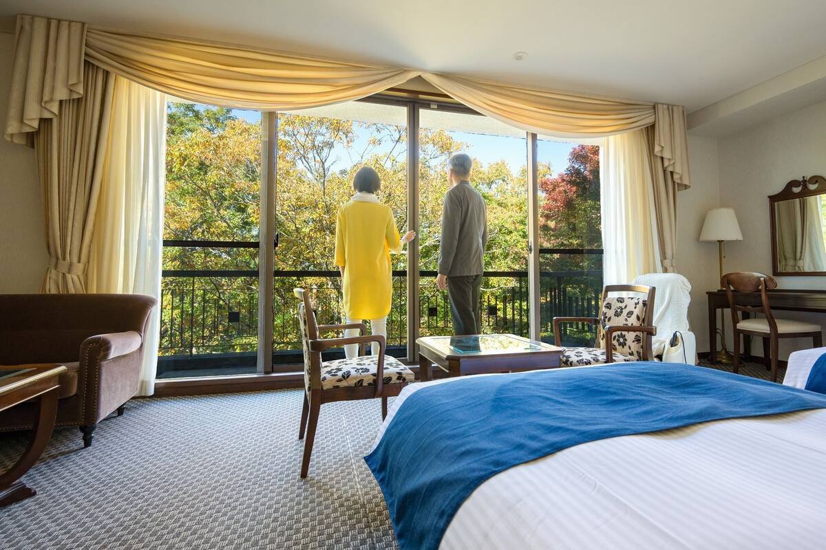 A hotel room with a large window, a bed, a desk, a chair, and a sofa.
