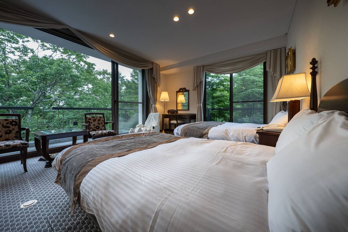 A hotel room with two beds, a desk, and a chair. There are large windows with curtains, a lamp, and a mirror on the wall.
