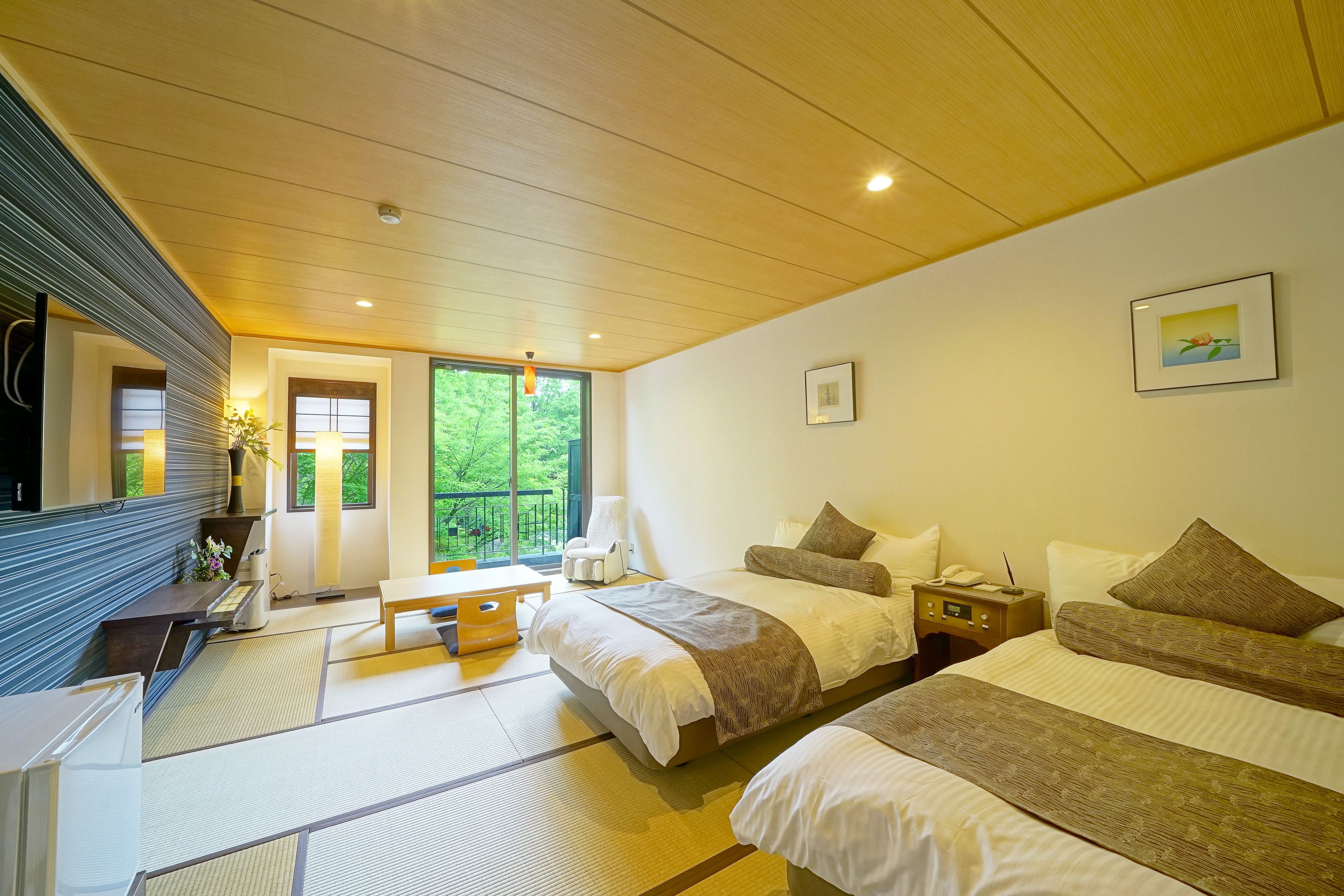 A hotel room with a large bed, a smaller bed, a desk, and a chair. There is a window with a view of greenery outside.