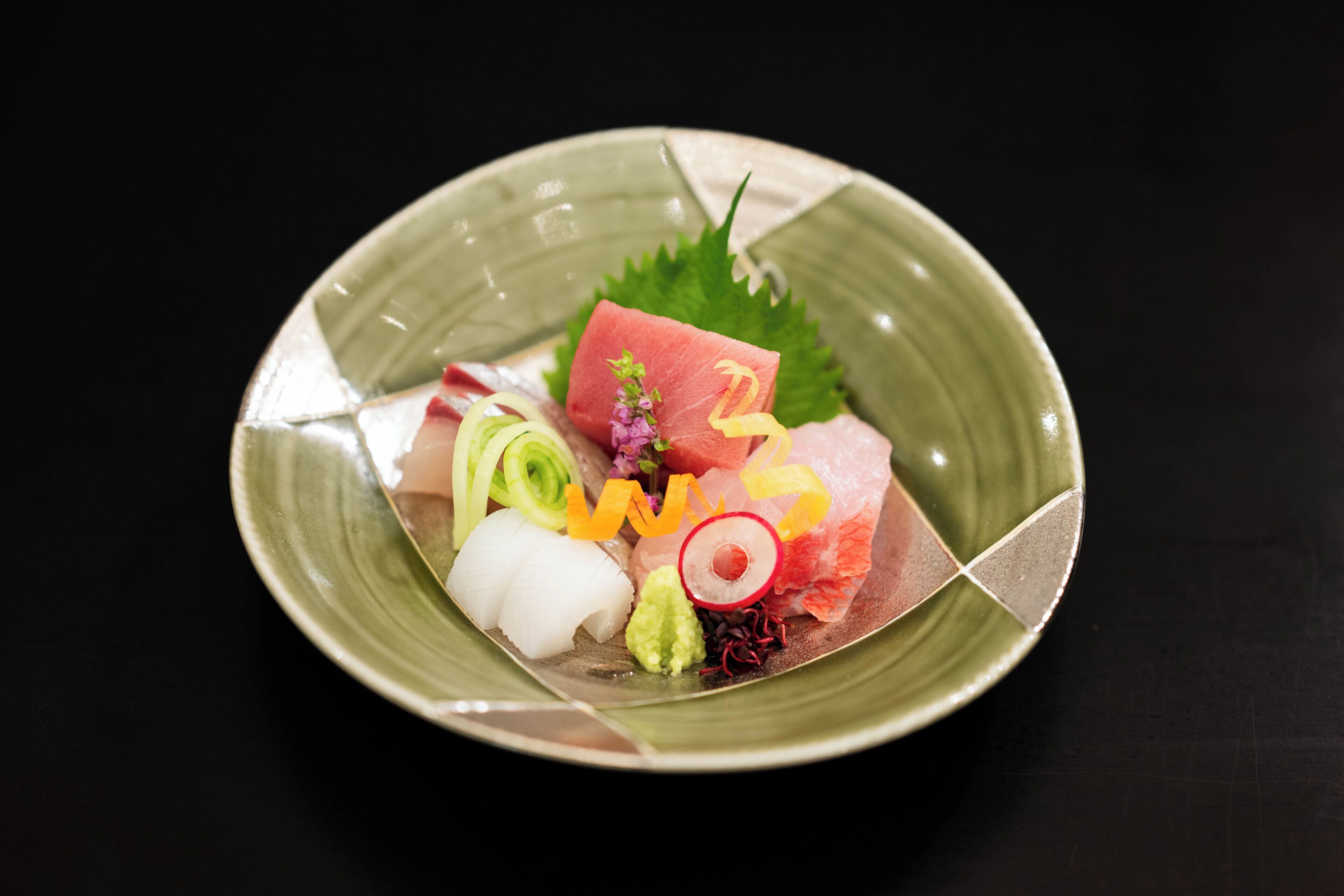 A green ceramic plate with various sashimi pieces, including salmon, tuna, and other seafood, garnished with green onions and a leaf.