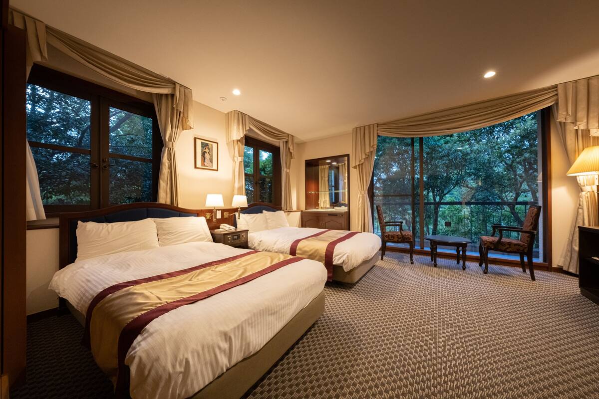 A hotel room with two beds, a desk, a chair, and large windows offering a view of trees.