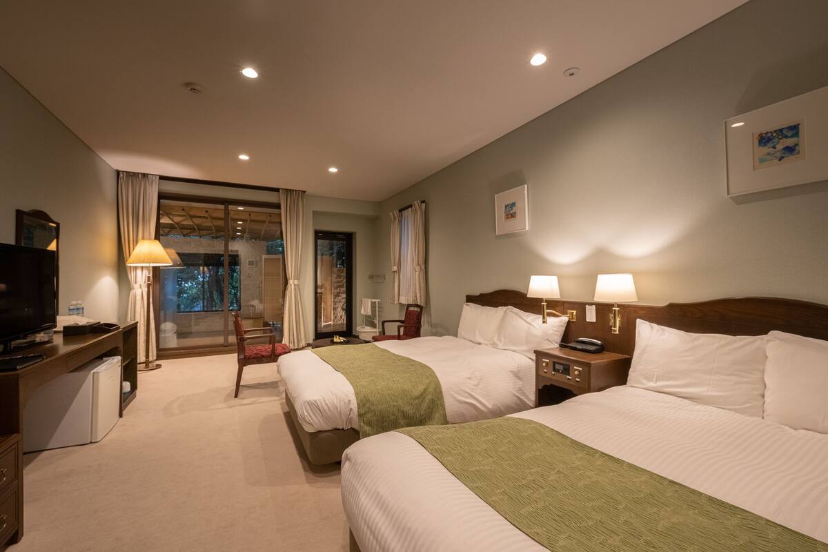 A hotel room with two beds, a desk with a television, a chair, and a view of the outdoors through a sliding door.