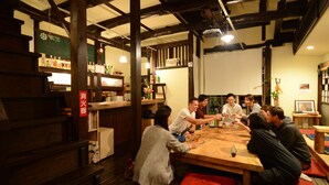 A group of people seated around a wooden table in a restaurant with wooden beams and a chalkboard menu. - Guesthouse Nin-jin - Hostel (Niigata)