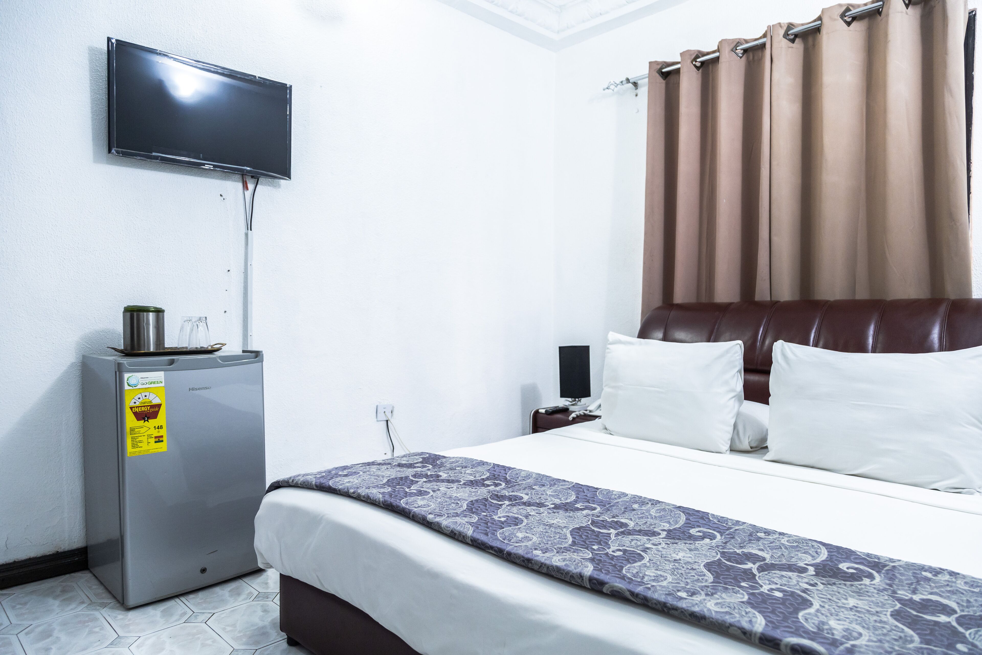 A hotel room with a bed, a television mounted on the wall, a small refrigerator, and a curtain.