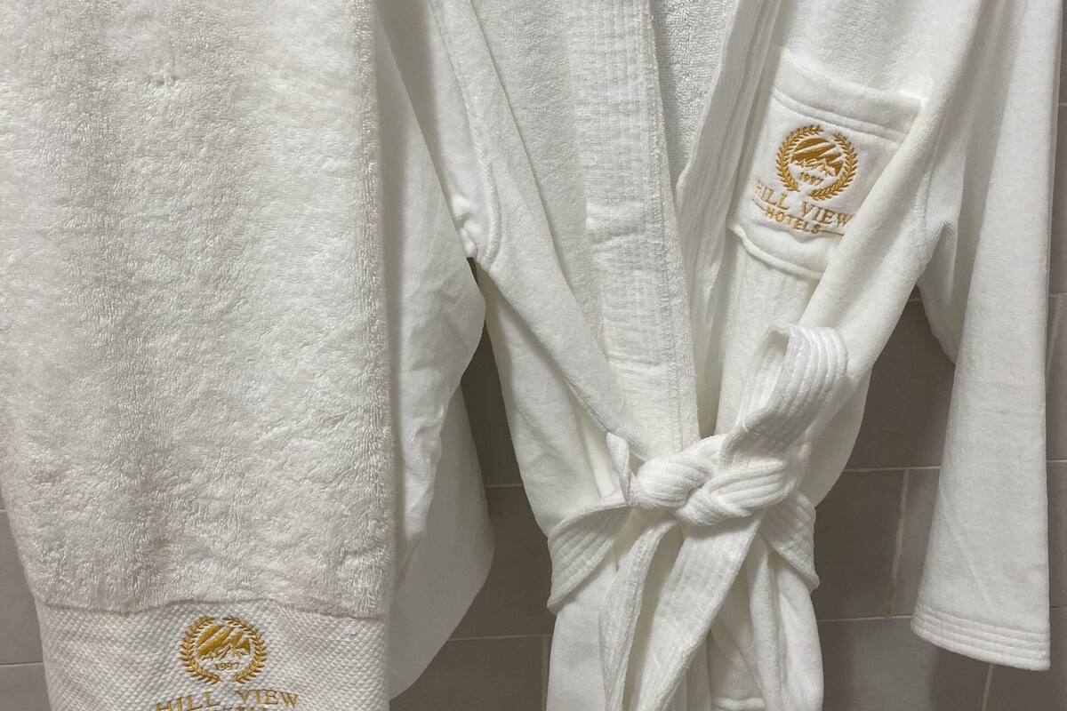 A white bathrobe with a gold embroidered logo and a towel hanging beside it.