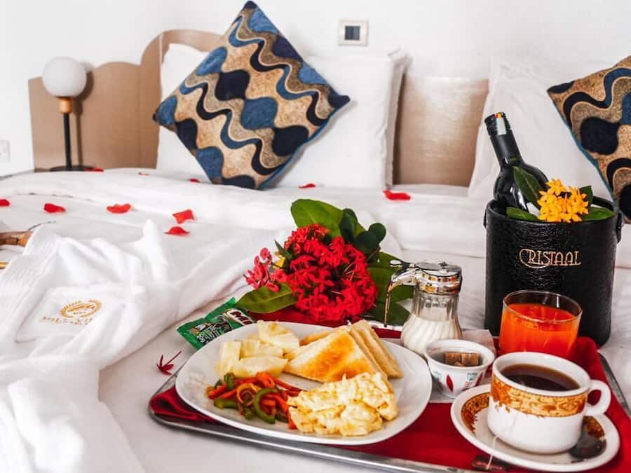 Un lit bien fait, recouvert d’une couverture à carreaux rouges et blancs, une bouteille de champagne Cristal, un verre de jus d’orange, une assiette d’œufs brouillés, des toasts et un petit bouquet de fleurs rouges.