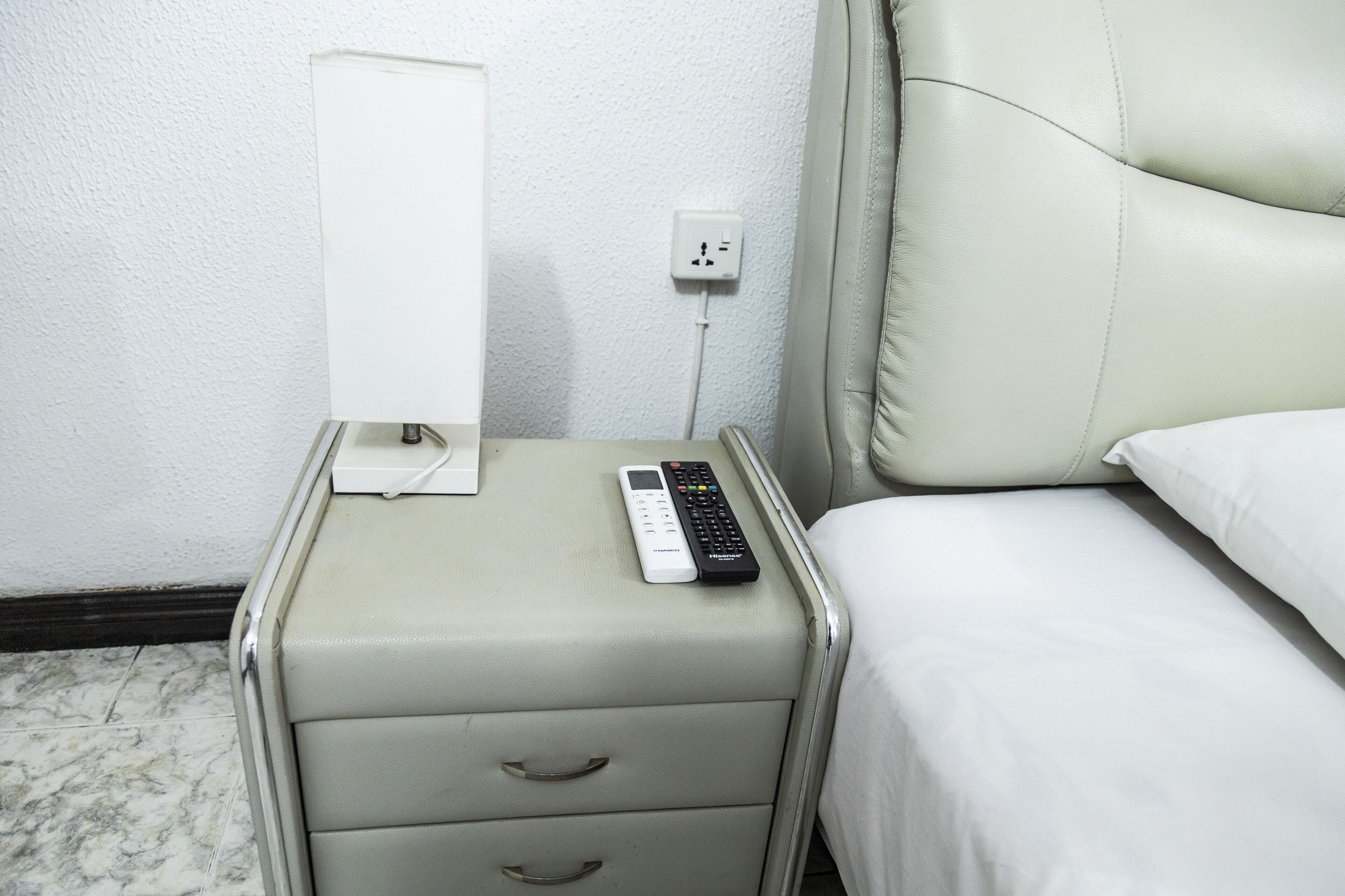 Habitación de hotel con cama, mesita de noche, control remoto, toma de corriente e interruptor de pared.