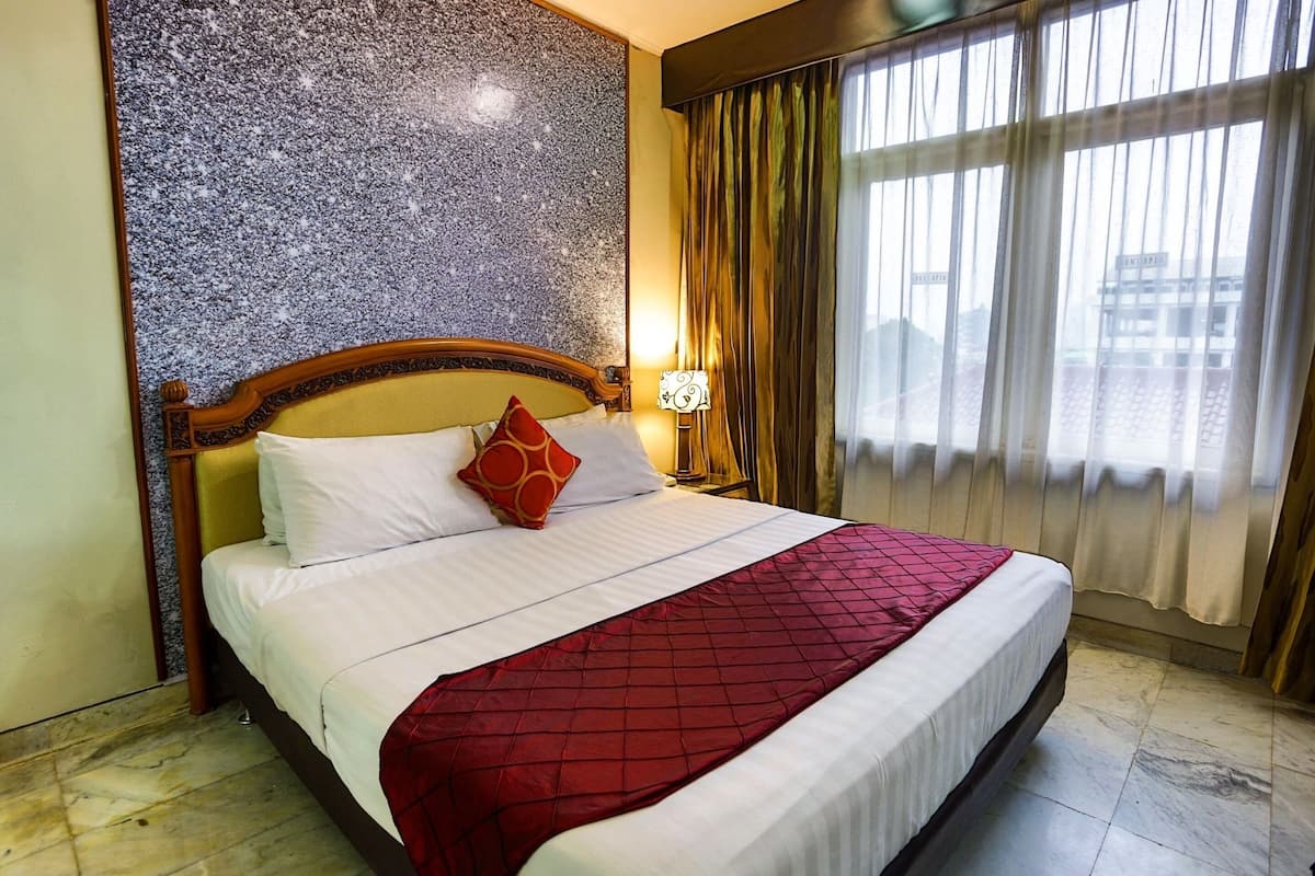 Habitación de hotel con una cama grande, cabecero y pared texturizada detrás, ventana con cortinas y mesita de noche con lámpara.