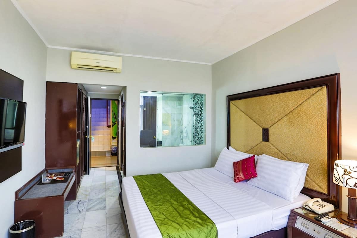 Habitación de hotel con una cama, una mesita de noche con lámpara, un teléfono y vista a un pasillo y otra habitación.
