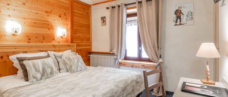 Una habitación con paneles de madera, una cama, un escritorio, una silla, un radiador y una ventana con cortinas.