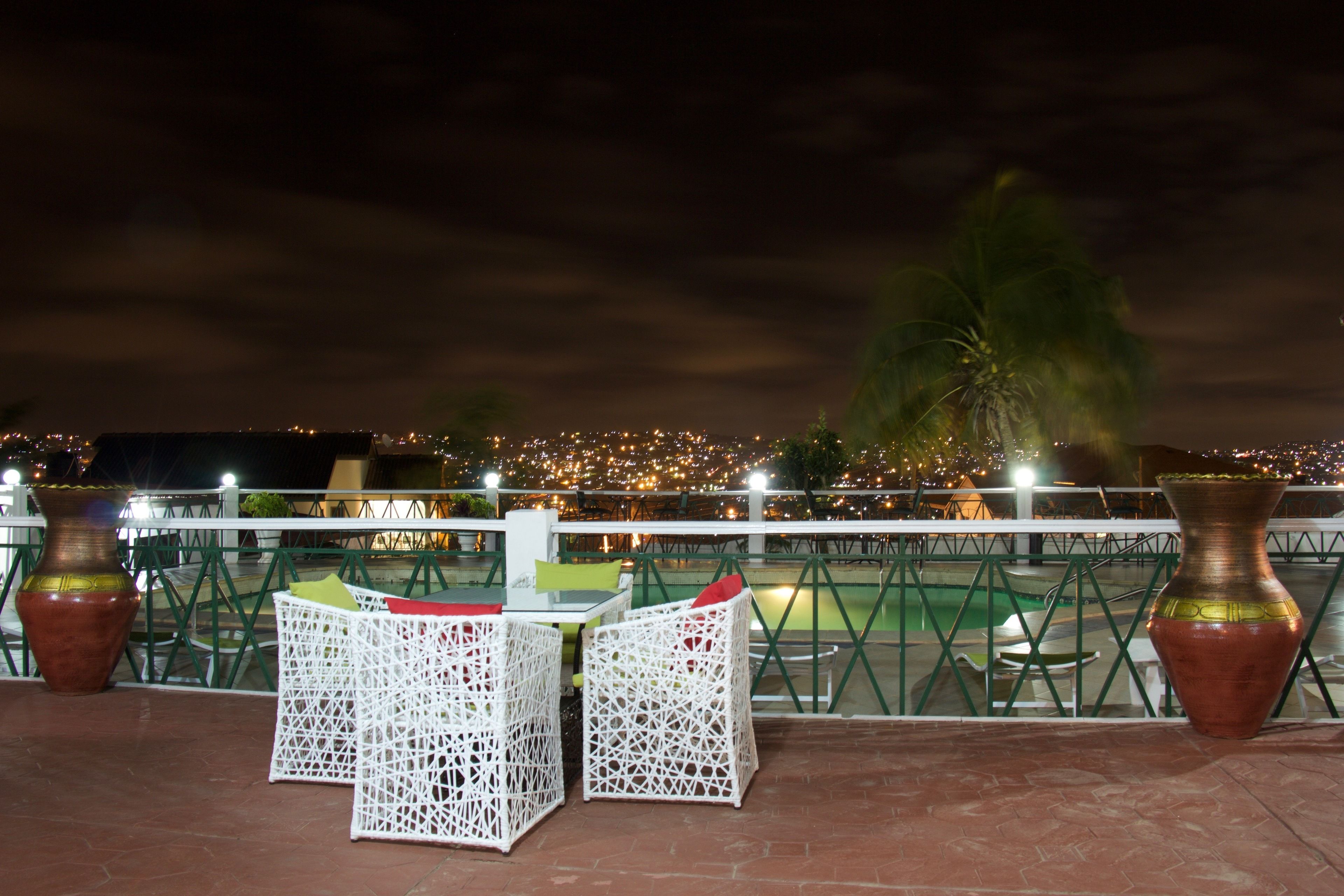 An outdoor seating area with white wicker chairs and a table, overlooking a cityscape at night.