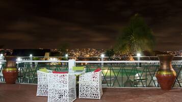 An outdoor seating area with white wicker chairs and a table, overlooking a cityscape at night.