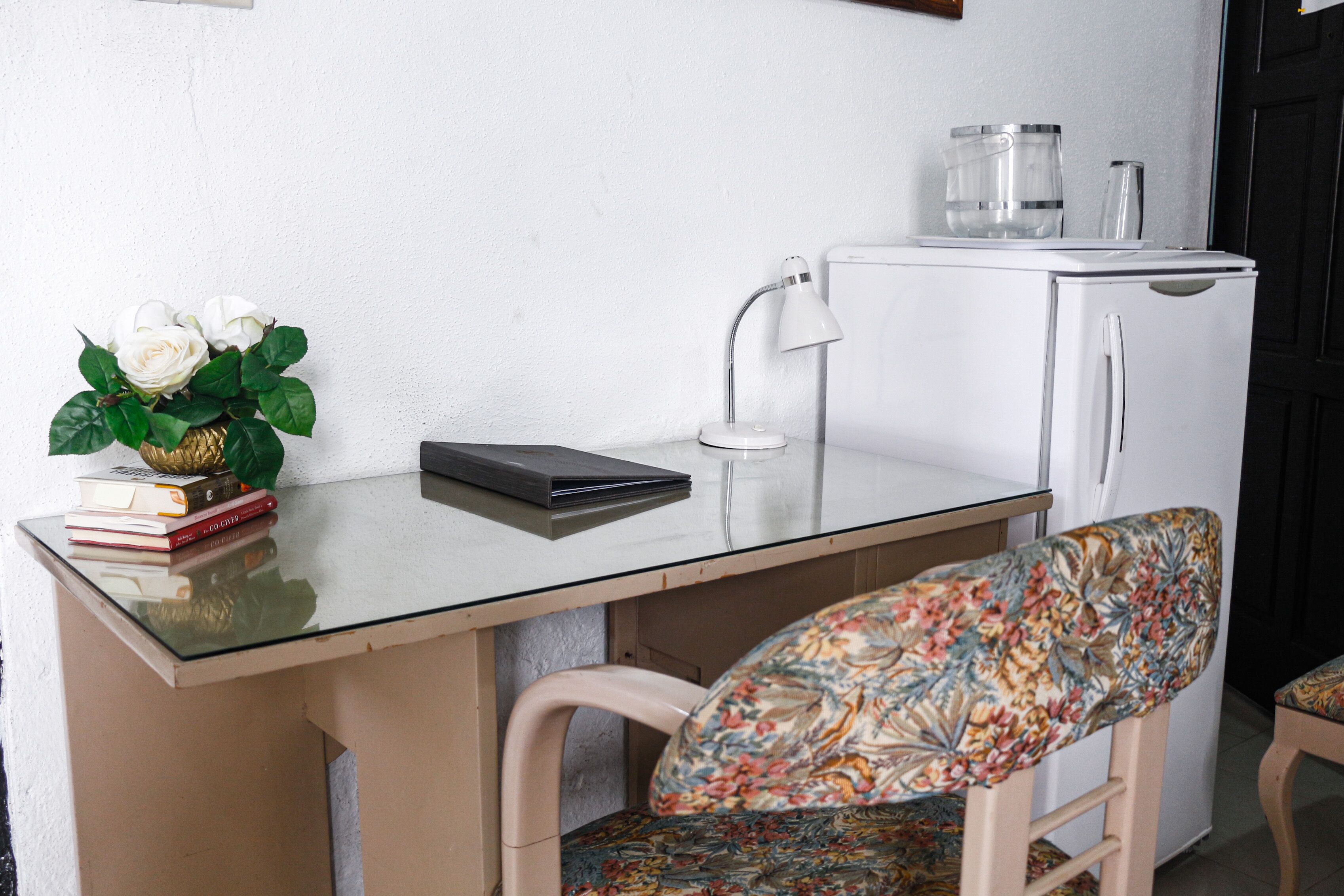 Una pequeña kitchenette con un refrigerador blanco, una lámpara blanca y un recipiente de vidrio encima. Hay una silla con un cojín floral y una mesa pequeña con superficie de vidrio.
