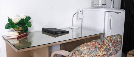 Una pequeña kitchenette con un refrigerador blanco, una lámpara blanca y un recipiente de vidrio encima. Hay una silla con un cojín floral y una mesa pequeña con superficie de vidrio.