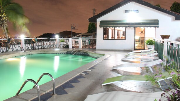 A swimming pool area with a covered seating area, a building with a sign, and a palm tree. - Hill View Hotel McCarthy Hills (Mallam)