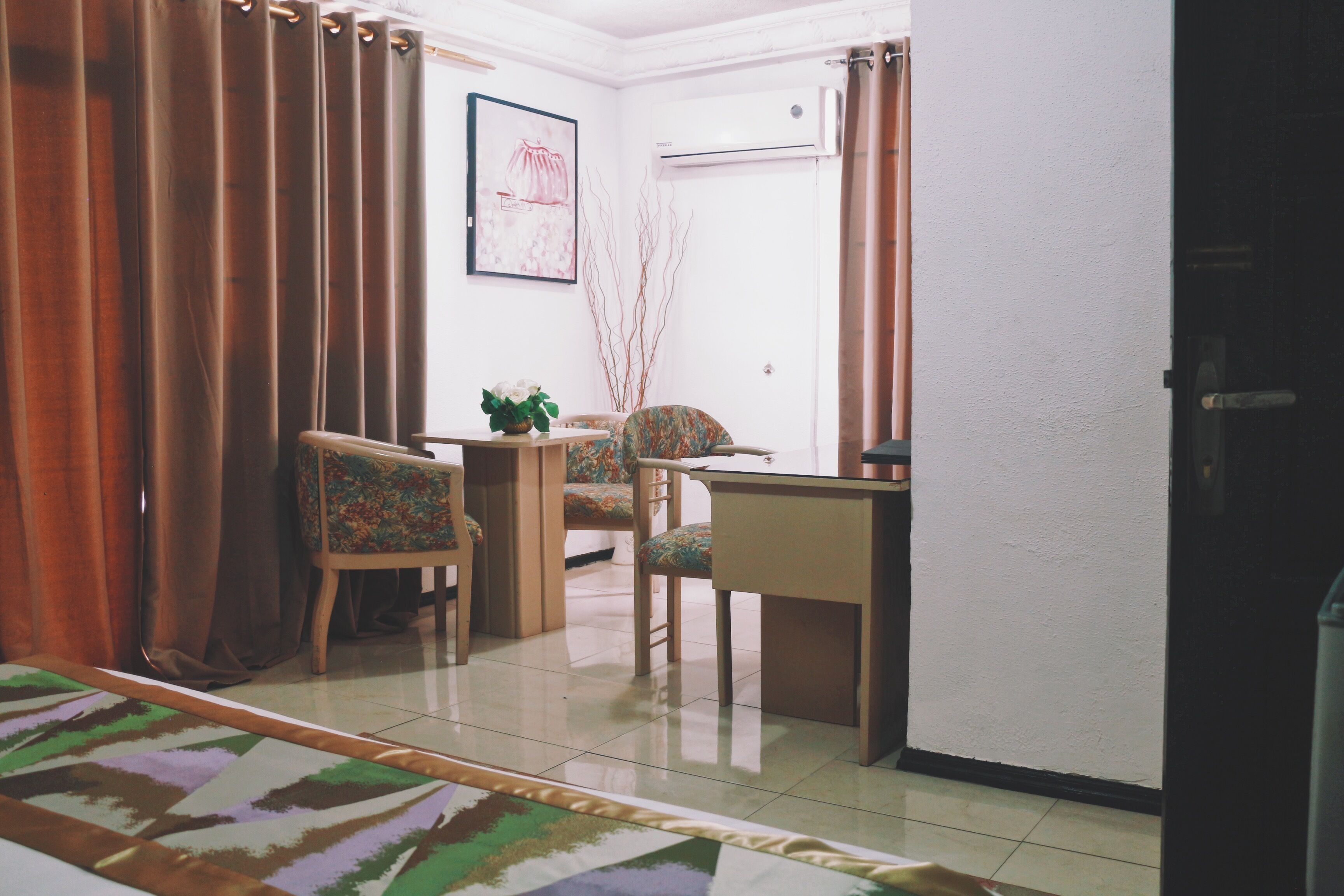 A room with a bed, two chairs, a small table, and a desk. There is a window with curtains, a wall-mounted air conditioner, and a framed picture on the wall.
