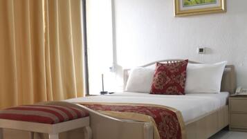 A hotel room with a bed, a bedside table, a nightstand, a window with curtains, a wall-mounted air conditioner, and a framed picture on the wall.